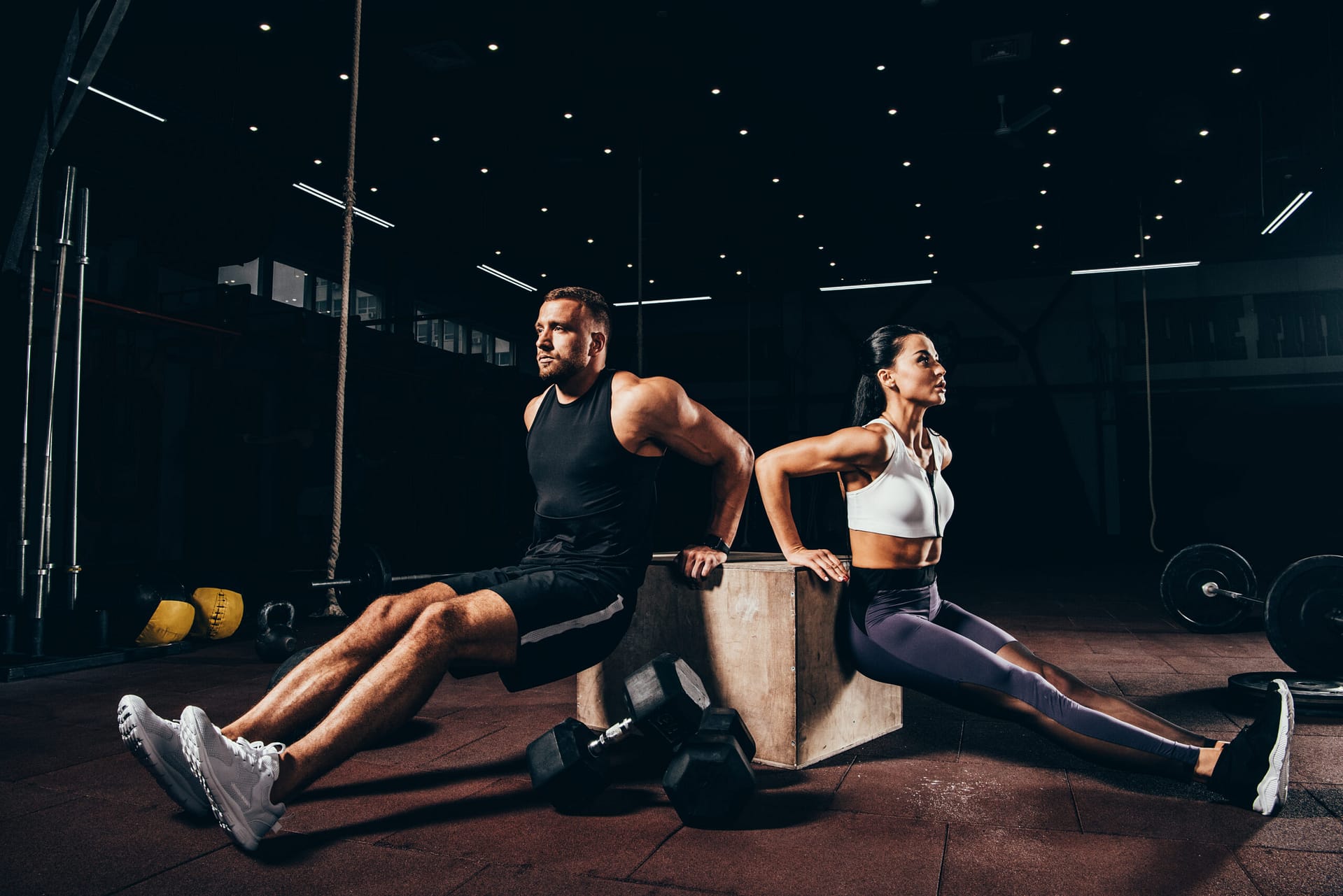 athletic sportsman and sportswoman exercising on cube together in dark gym